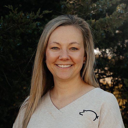 A woman with long blonde hair smiles at the camera, standing outdoors in front of green trees. She is wearing a light-colored top with a simple embroidered design.
